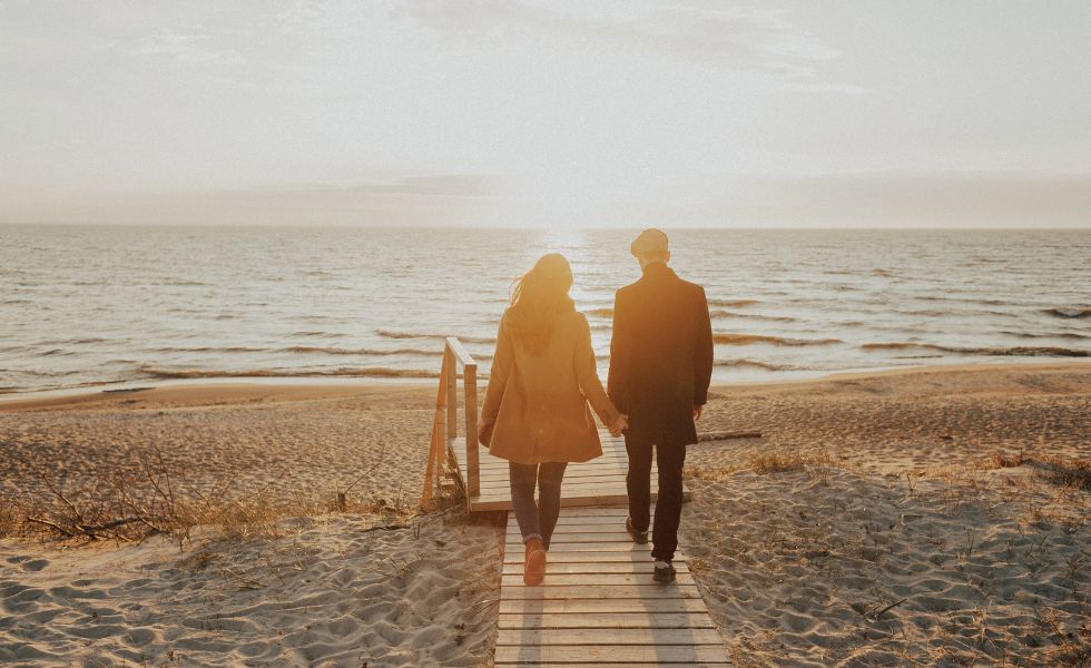 Para, która idzie po kładce na plaży za rękę na tle morza, przy zachodzącym słońcu